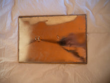Load image into Gallery viewer, a shot of the back of the mirror. It&#39;s plain brass, with a small hook for hanging and the signature of the maker.
